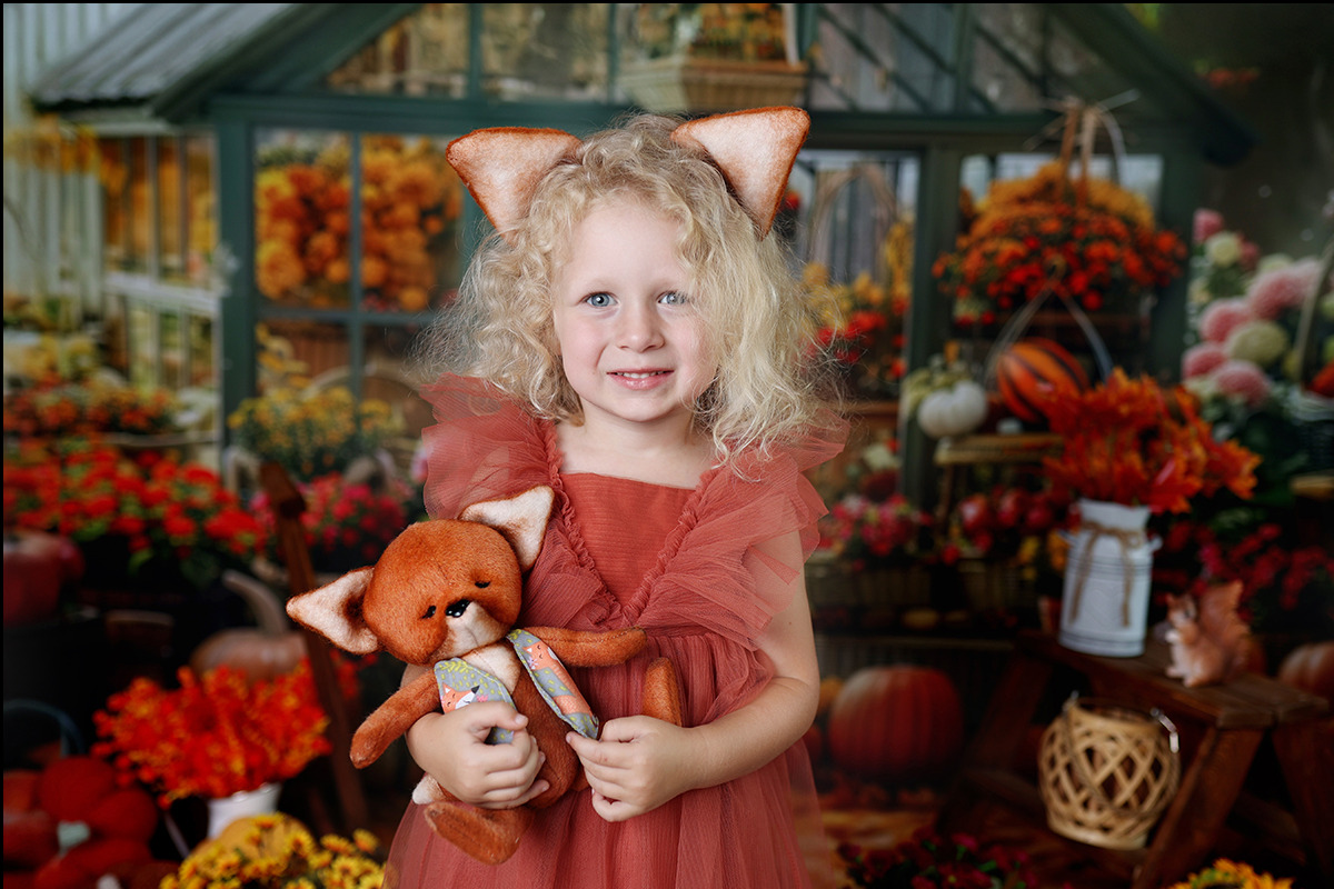 Uśmiechnięta dziewczynka z blond lokami i lisimi uszkami trzyma pluszowego liska na tle jesiennej dekoracji z dyniami i kwiatami w oranżerii.