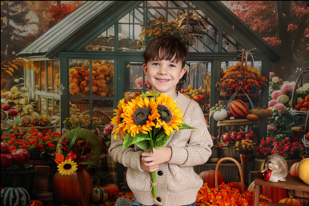 Uśmiechnięty chłopiec w beżowym swetrze trzyma bukiet słoneczników na tle dekoracji jesiennych, w tym dyni, kwiatów i miniaturowej szklarni.