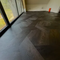 Ukończona podłoga z ciemnych, drewnianych paneli ułożonych we wzór jodełki francuskiej w jasnym pokoju z dużym oknem.