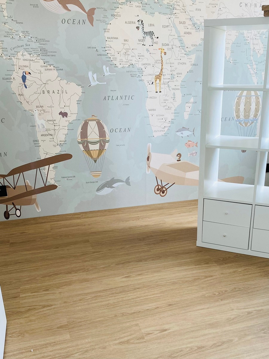 Jasna podłoga drewniana w pokoju dziecięcym z tapetą przedstawiającą mapę świata z motywami podróżniczymi, takimi jak balony i samoloty, oraz biały regał z szufladami.