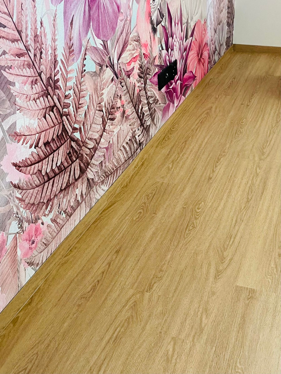 Jasne panele podłogowe ułożone wzdłuż ściany z fototapetą w kwiaty i paprocie, widoczny fragment gniazdka elektrycznego.