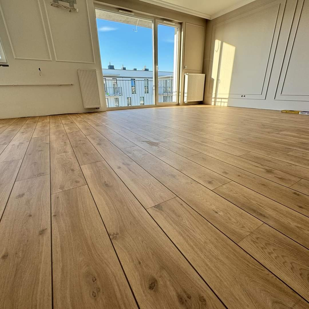 Jasne panele podłogowe ułożone w przestronnym pokoju z dużym oknem balkonowym i dekoracyjnymi listwami ściennymi, widok na budynek z balkonami.