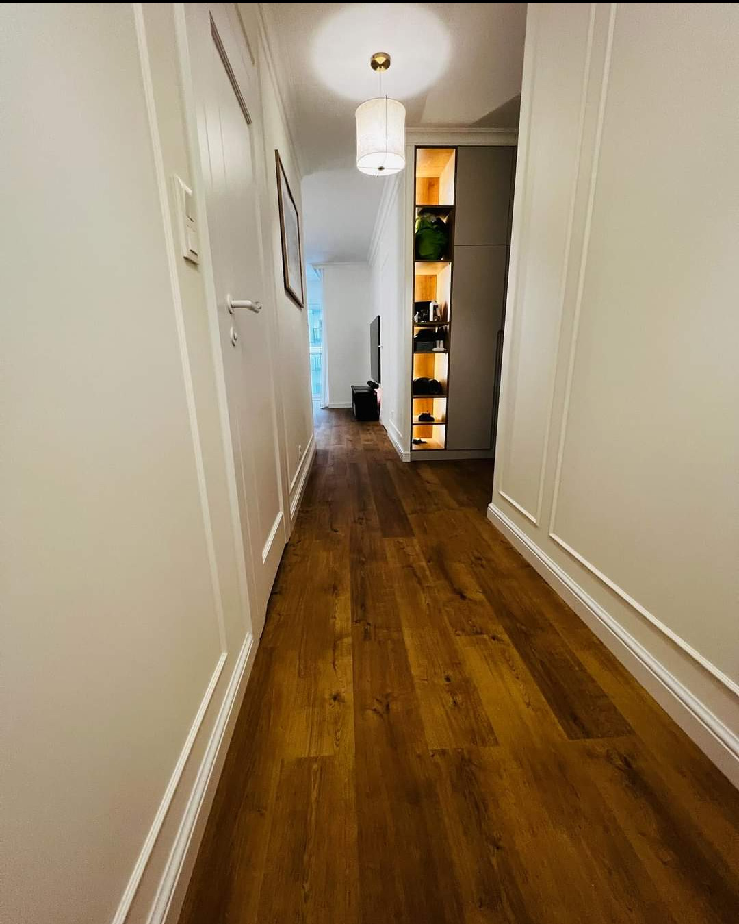 Korytarz z nową, ciemną podłogą imitującą drewno, z wbudowaną szafą z podświetlanymi półkami i białymi ścianami z dekoracyjnymi listwami.