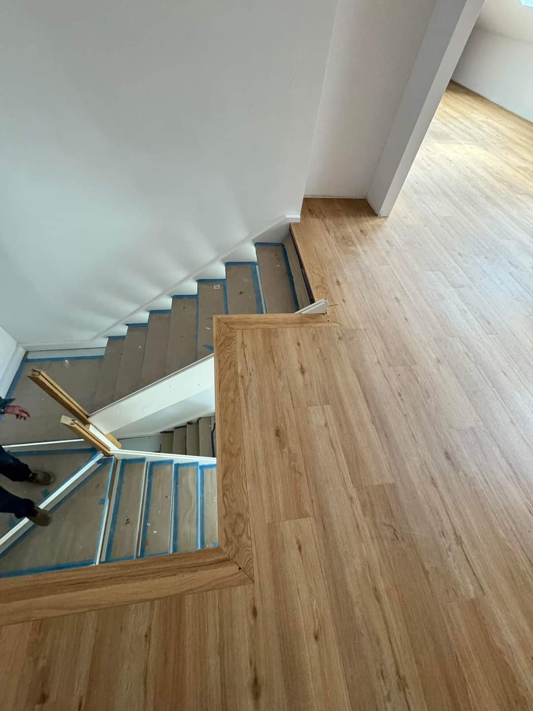 Montaż drewnianej podłogi i schodów, widoczne zabezpieczone schody z kartonem i niebieską taśmą, oraz fragment podłogi z jasnego drewna.
