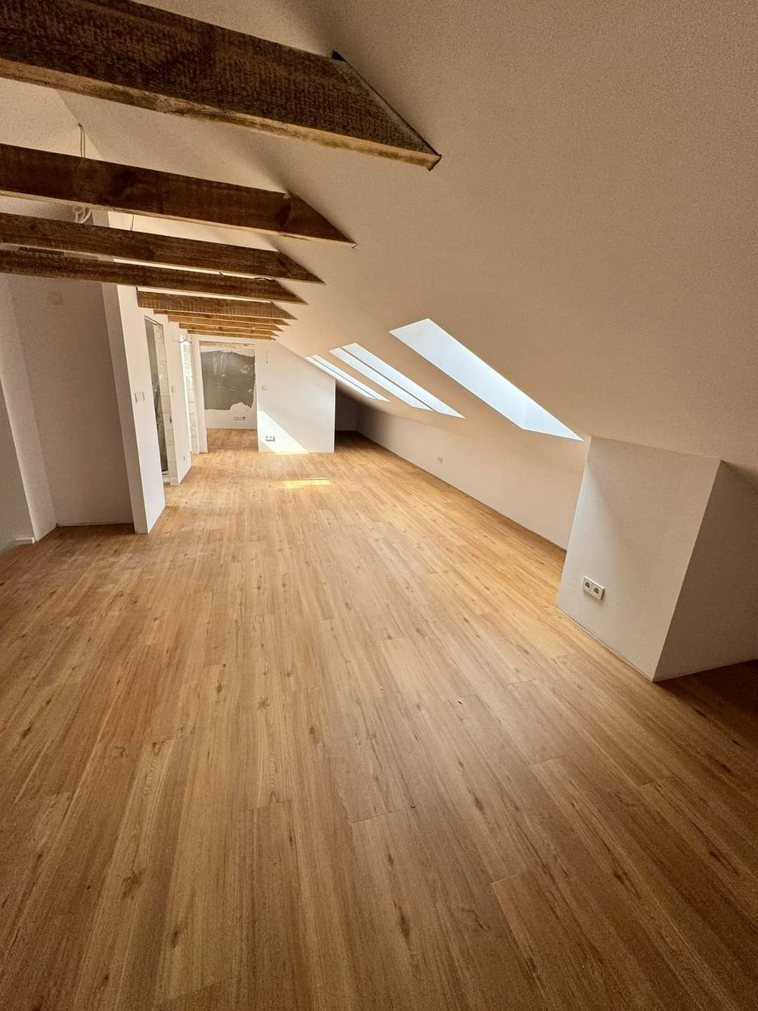 Jasna, drewnopodobna podłoga w nowym, przestronnym pokoju na poddaszu z belkami stropowymi i oknami dachowymi.
