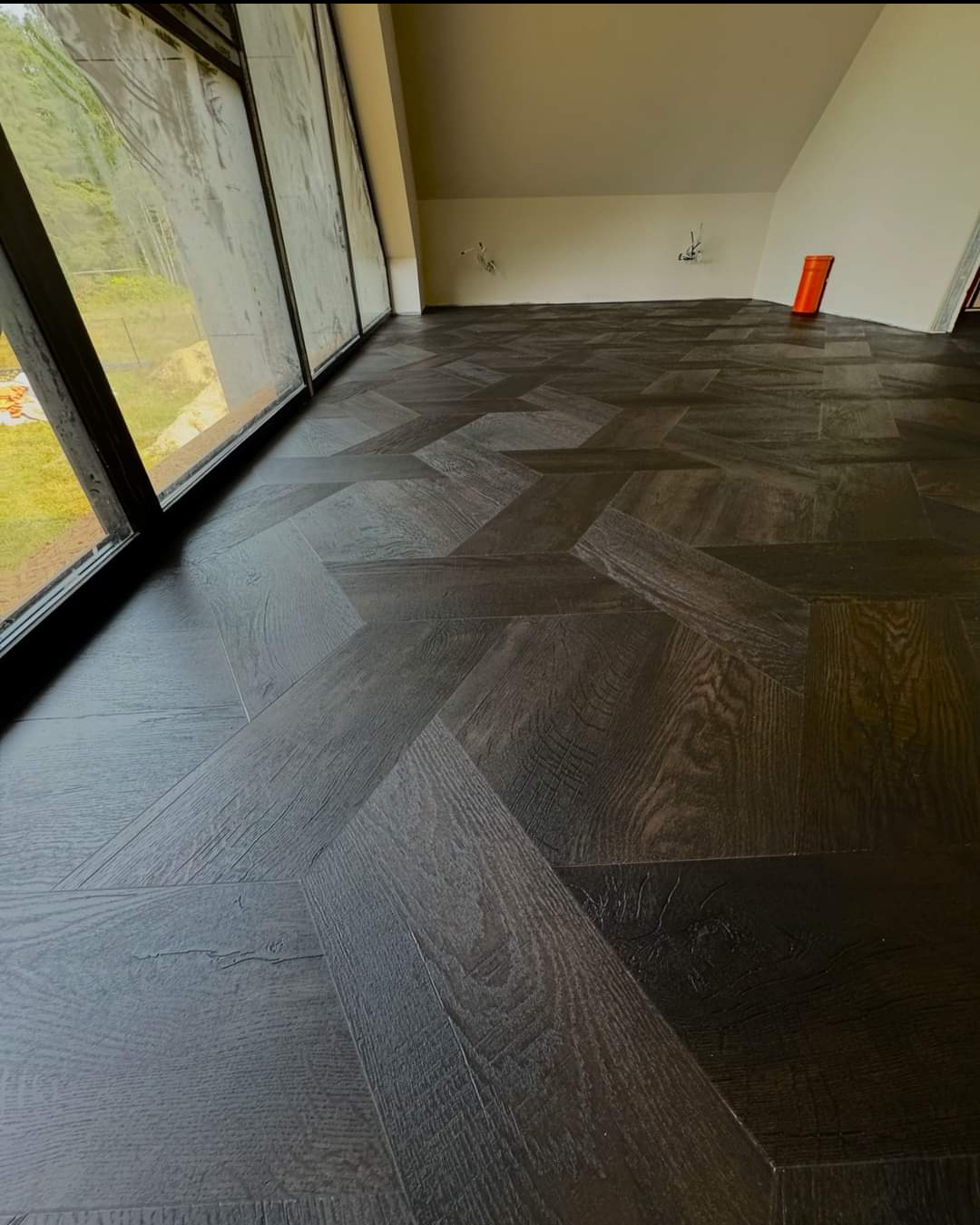 Ukończona podłoga z ciemnych, drewnianych paneli ułożonych we wzór jodełki francuskiej w jasnym pokoju z dużym oknem.