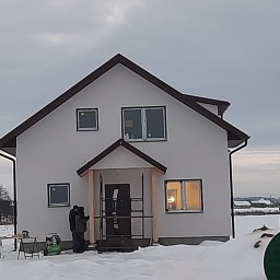 CONSTRUCTION Firma Budowlana Andrzej Hamelusz - Niedokończony biały dom z brązowym dachem w zimowym krajobrazie; widoczny fragment budowanego ganku i sprzęt budowlany na zewnątrz.