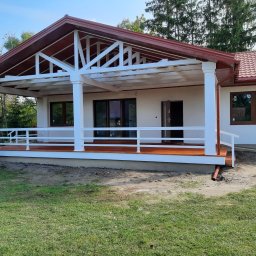 CONSTRUCTION Firma Budowlana Andrzej Hamelusz - Gotowy dom szkieletowy z zadaszonym tarasem, białe kolumny podtrzymujące dach, brązowa dachówka i stolarka okienna, teren wokół domu częściowo zagospodarowany.