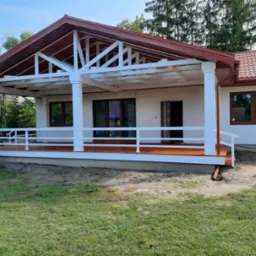 Gotowy dom szkieletowy z zadaszonym tarasem, białe kolumny podtrzymujące dach, brązowa dachówka i stolarka okienna, teren wokół domu częściowo zagospodarowany.