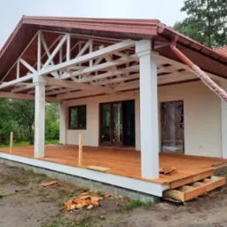 Budowa domu: widok na drewniany taras z białymi kolumnami i konstrukcją dachu, z elementami wykończeniowymi i fragmentami cegieł na ziemi.