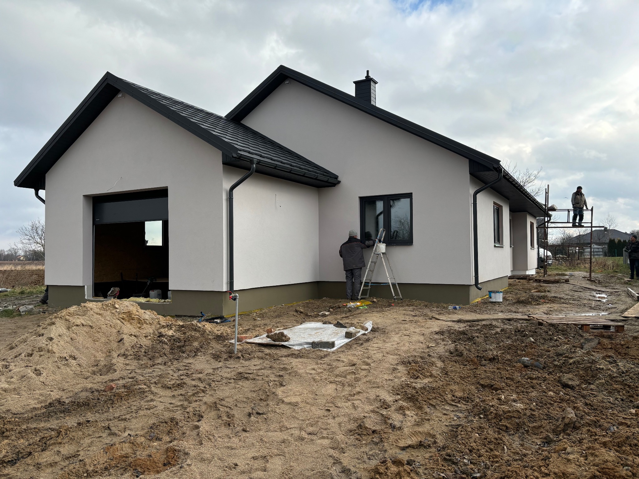 Nowoczesny parterowy dom z garażem w trakcie wykończenia elewacji; pracownik maluje ramę okienną, drugi pracuje na rusztowaniu.