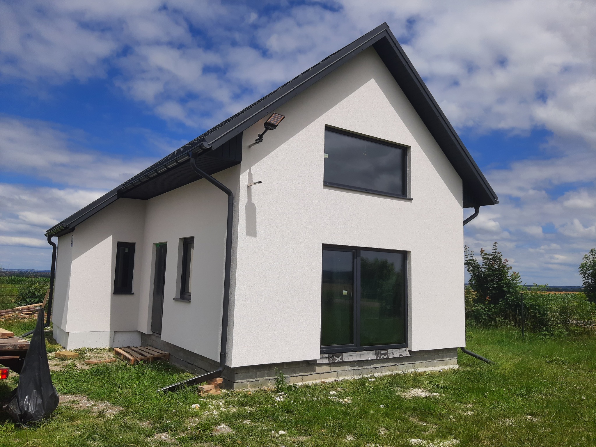 Nowoczesny, biały budynek z ciemną stolarką okienną i dachem na tle błękitnego nieba z chmurami. Widoczny fragment dachu z rynnami, lampa solarna na elewacji i paleta przy ścianie.