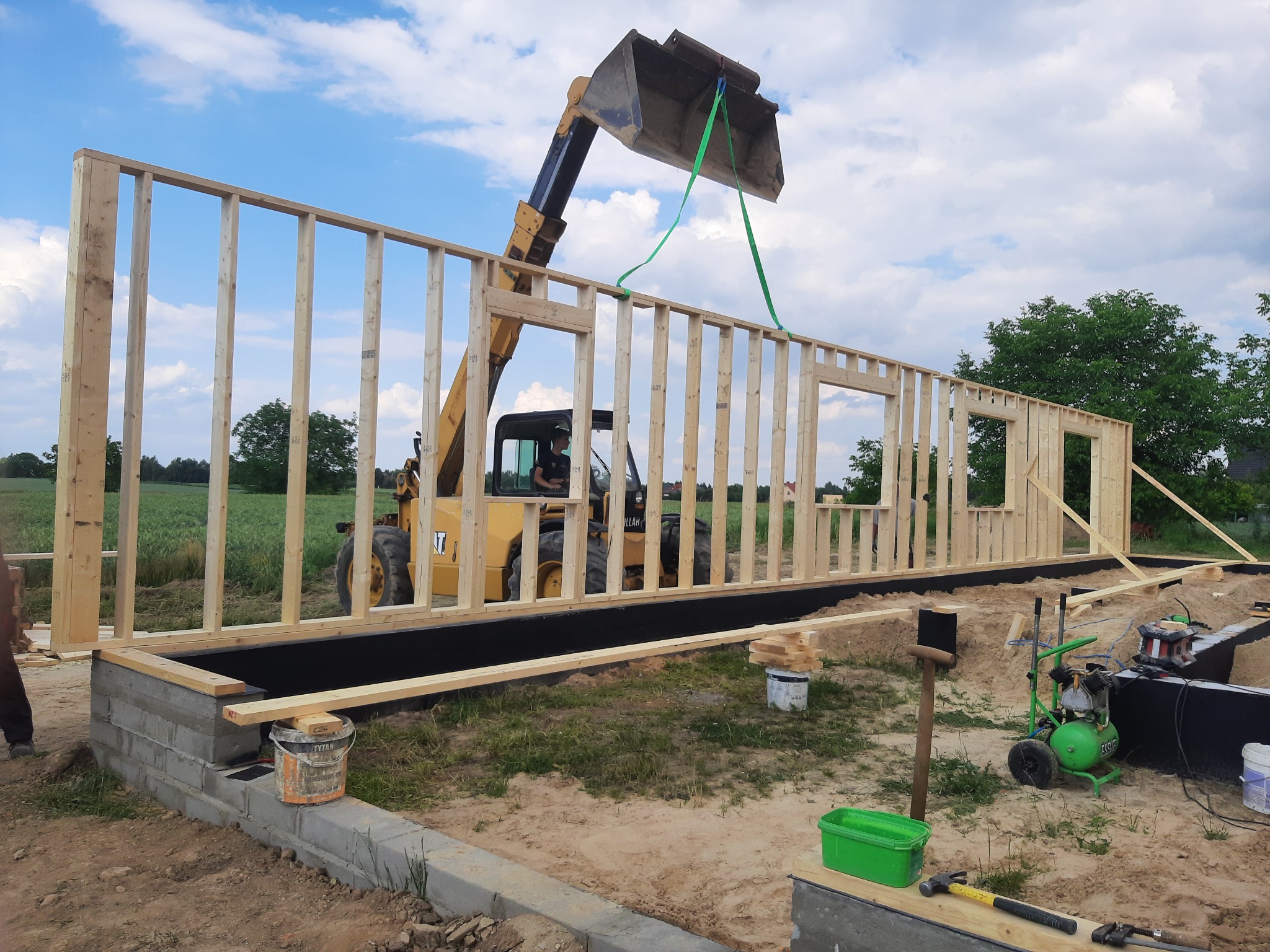 Montaż ściany szkieletowej domu za pomocą ładowarki teleskopowej na fundamencie, widoczny kompresor i narzędzia budowlane