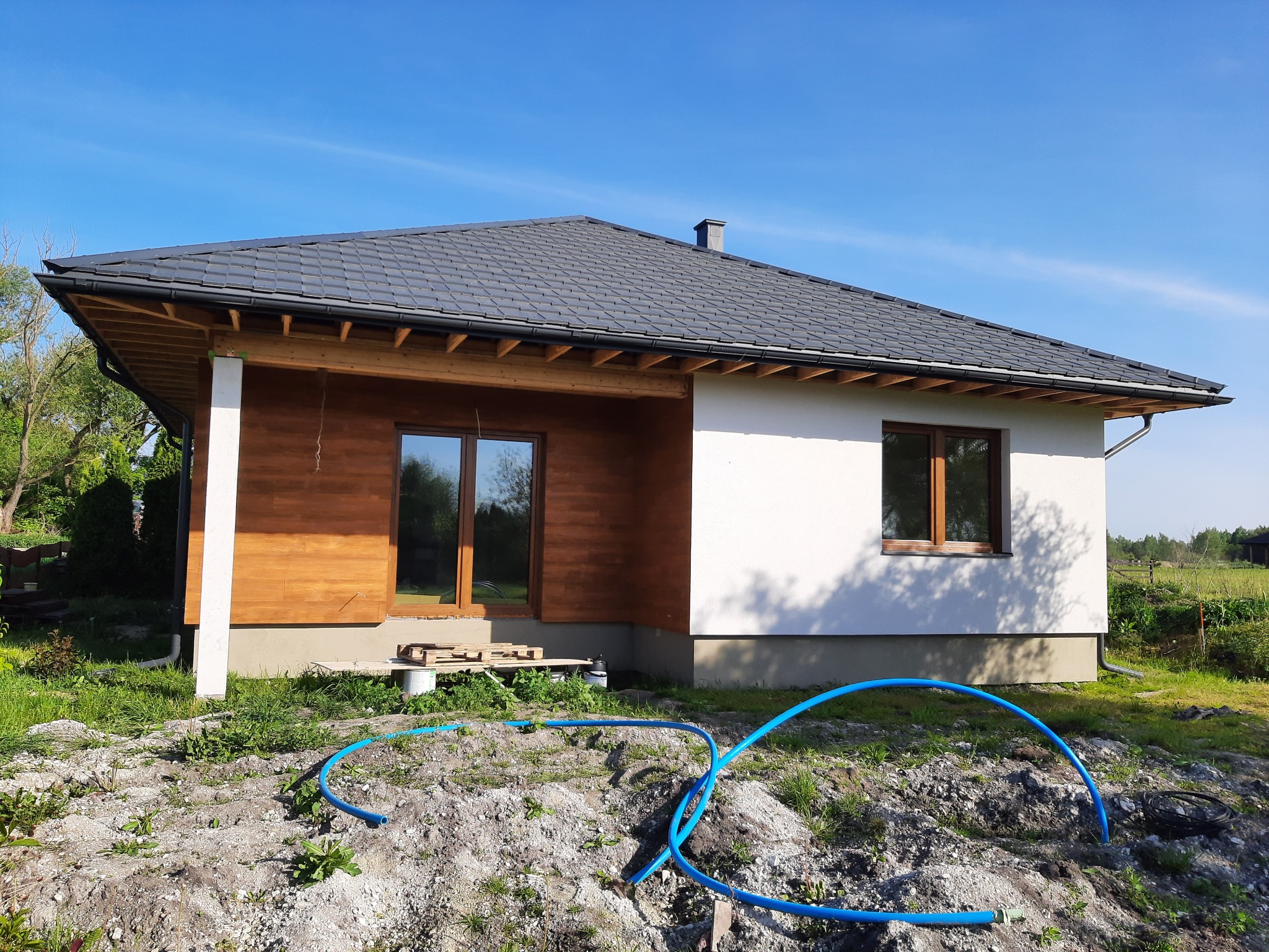 Surowy stan jednopiętrowego domu z drewnianą elewacją i szarą dachówką, niebieskie rury na ziemi.