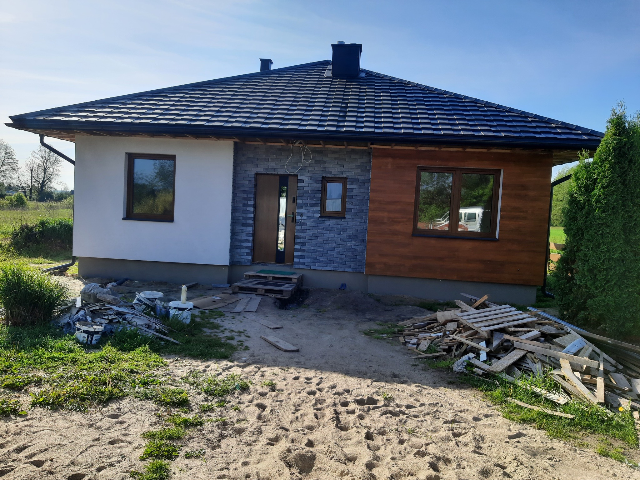 Niedokończony dom z elewacją w trzech stylach: biały tynk, szara cegła i drewniana boazeria, widoczne materiały budowlane i palety przed wejściem.