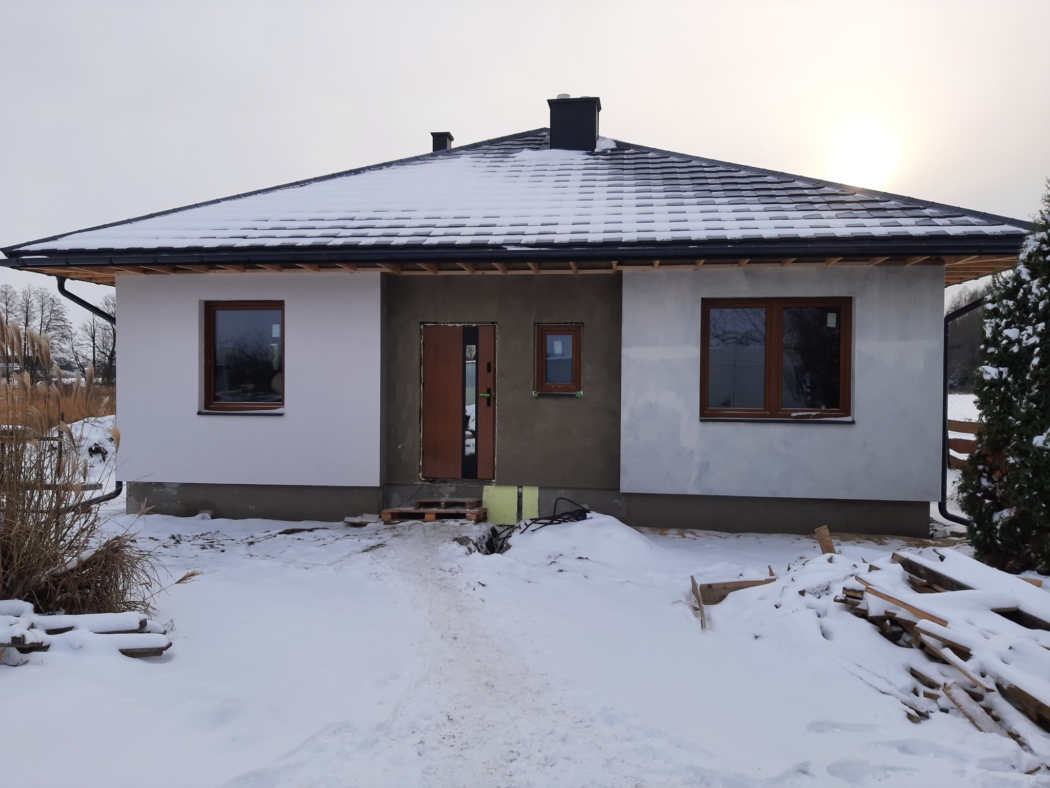 Surowy stan budynku jednorodzinnego z dachem pokrytym śniegiem, widoczne okna z brązowymi ramami i nieotynkowana elewacja przy wejściu.