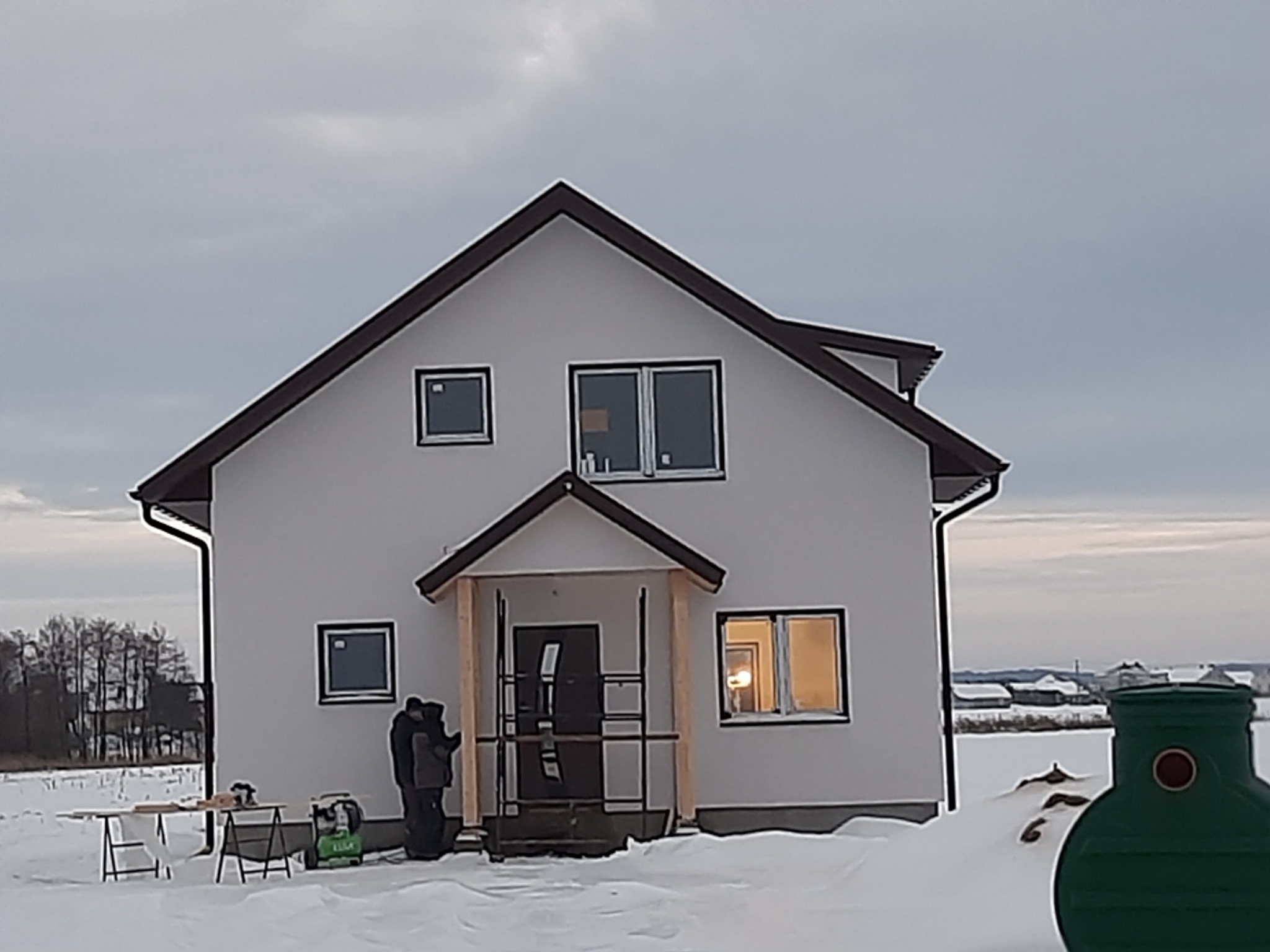 Niedokończony biały dom z brązowym dachem w zimowym krajobrazie; widoczny fragment budowanego ganku i sprzęt budowlany na zewnątrz.