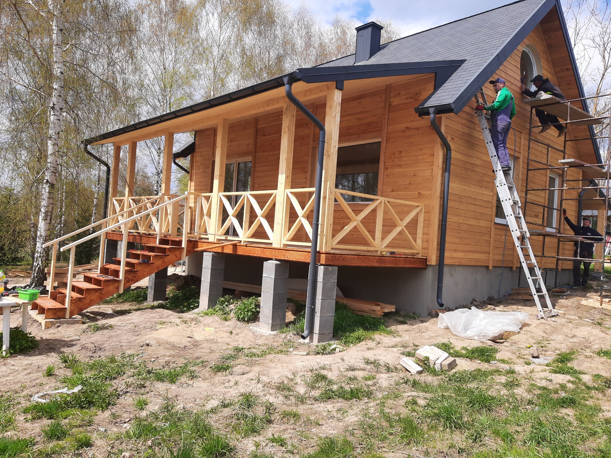 Wznoszenie drewnianego domu z elementami szkieletowymi, widoczne rusztowania, pracownicy montujący elementy wykończeniowe dachu i orynnowania, schody wejściowe z drewnianą balustradą, dom...