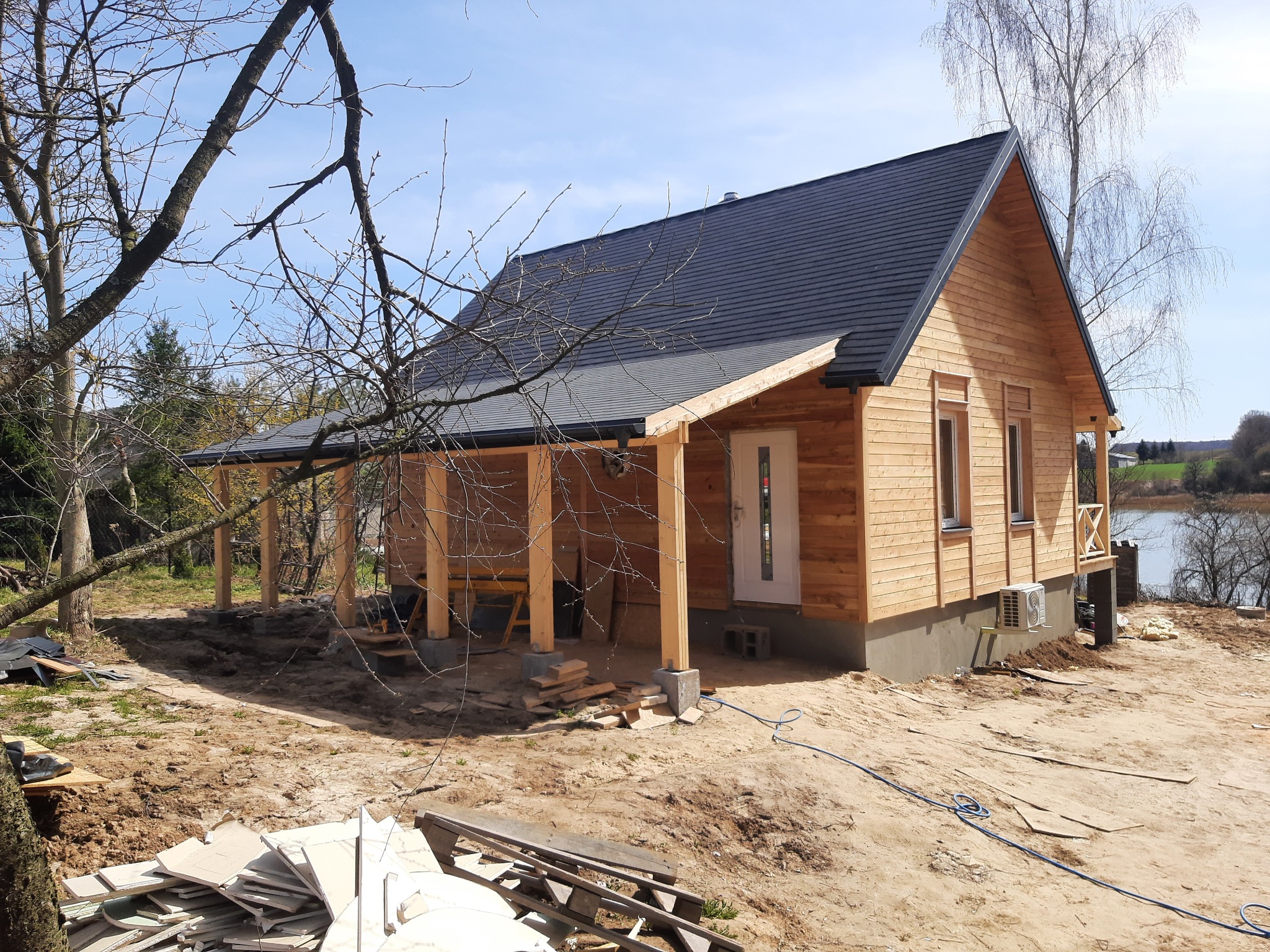 Surowy stan budowy parterowego domu z drewna z ciemnym dachem, widoczne drewniane kolumny ganku, teren wokół domu w trakcie prac ziemnych, w tle jezioro i drzewa.