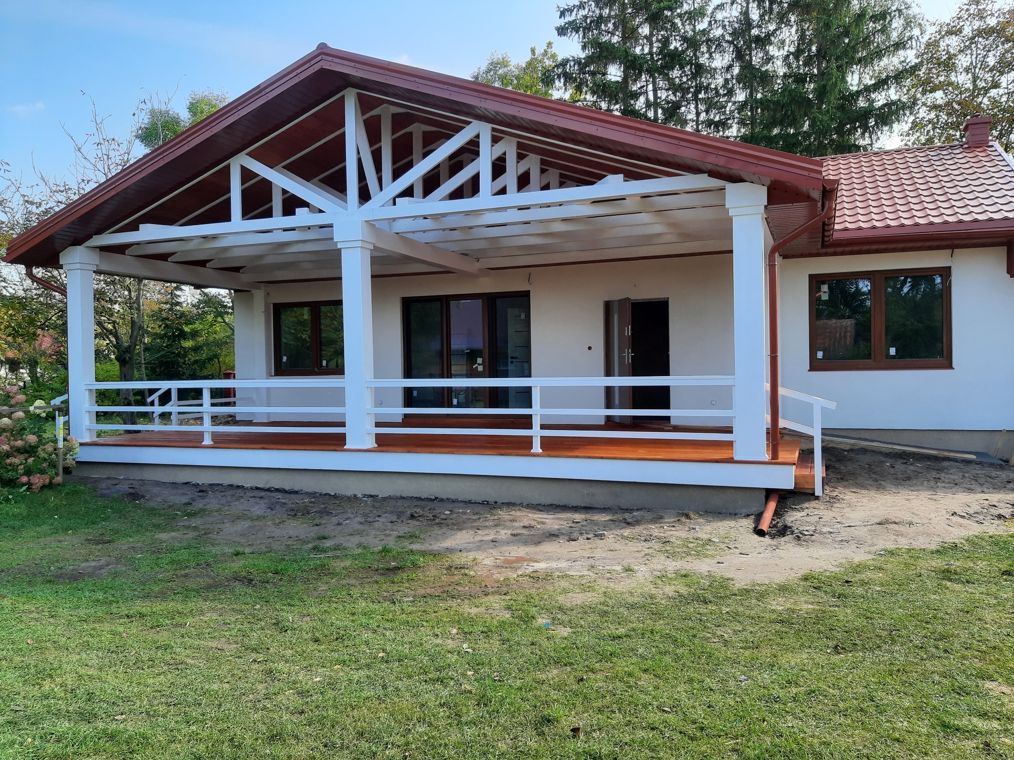 Gotowy dom szkieletowy z zadaszonym tarasem, białe kolumny podtrzymujące dach, brązowa dachówka i stolarka okienna, teren wokół domu częściowo zagospodarowany.