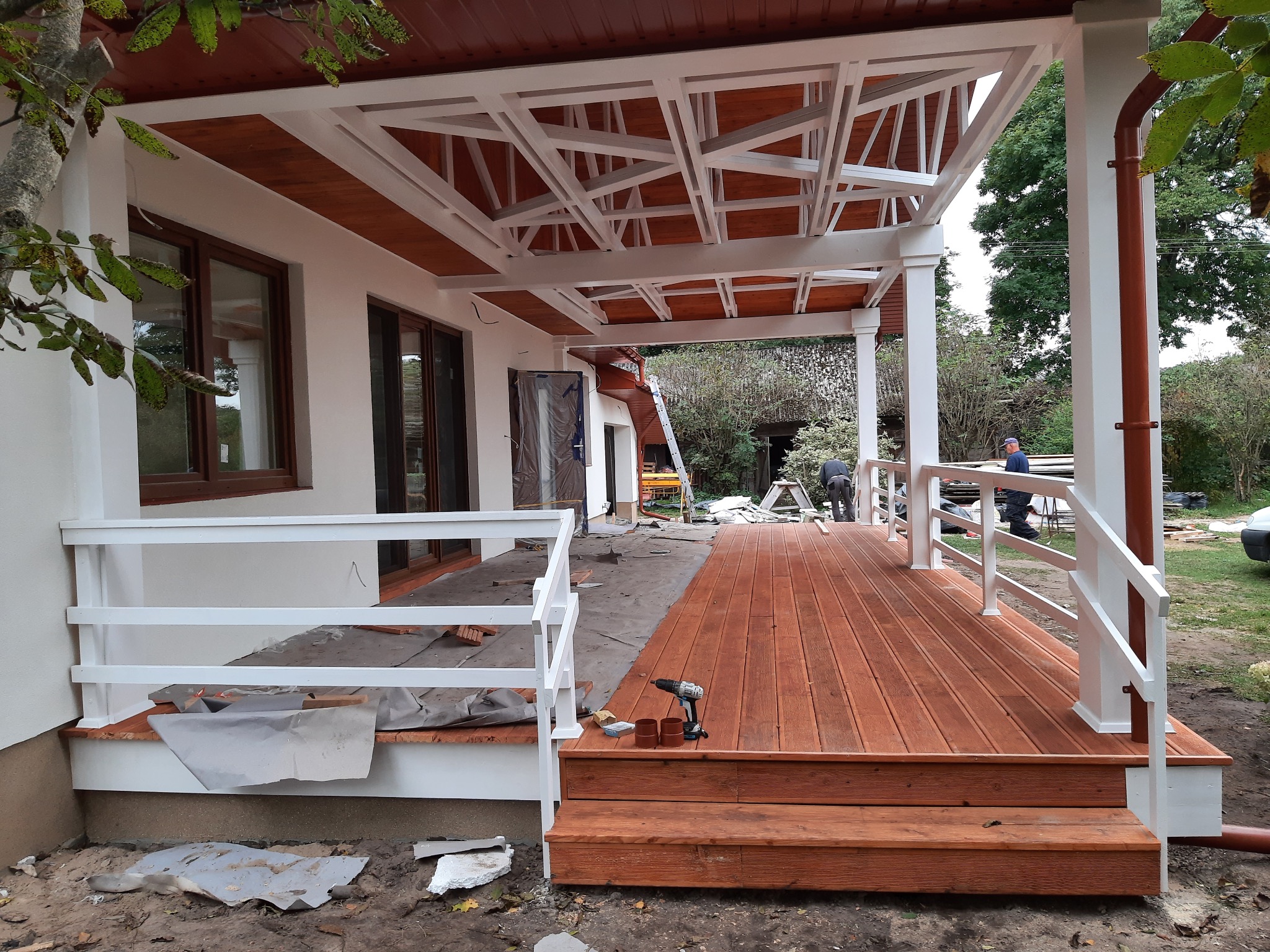 Zadaszony taras z drewnianą podłogą w trakcie budowy, widoczna wkrętarka na schodach, częściowo odkryte podłoże i pracownicy w tle.