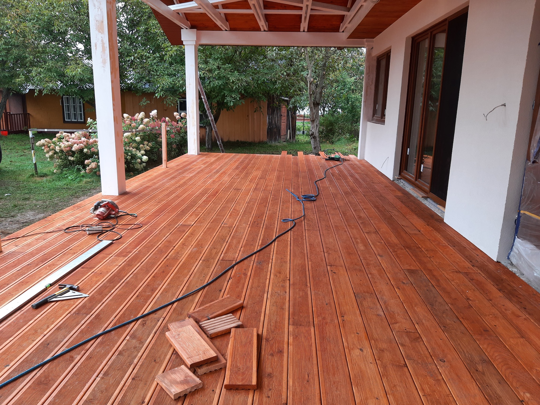 Montaż drewnianego tarasu przy domu szkieletowym, widoczne deski, narzędzia i piła elektryczna na świeżo ułożonej podłodze tarasowej.