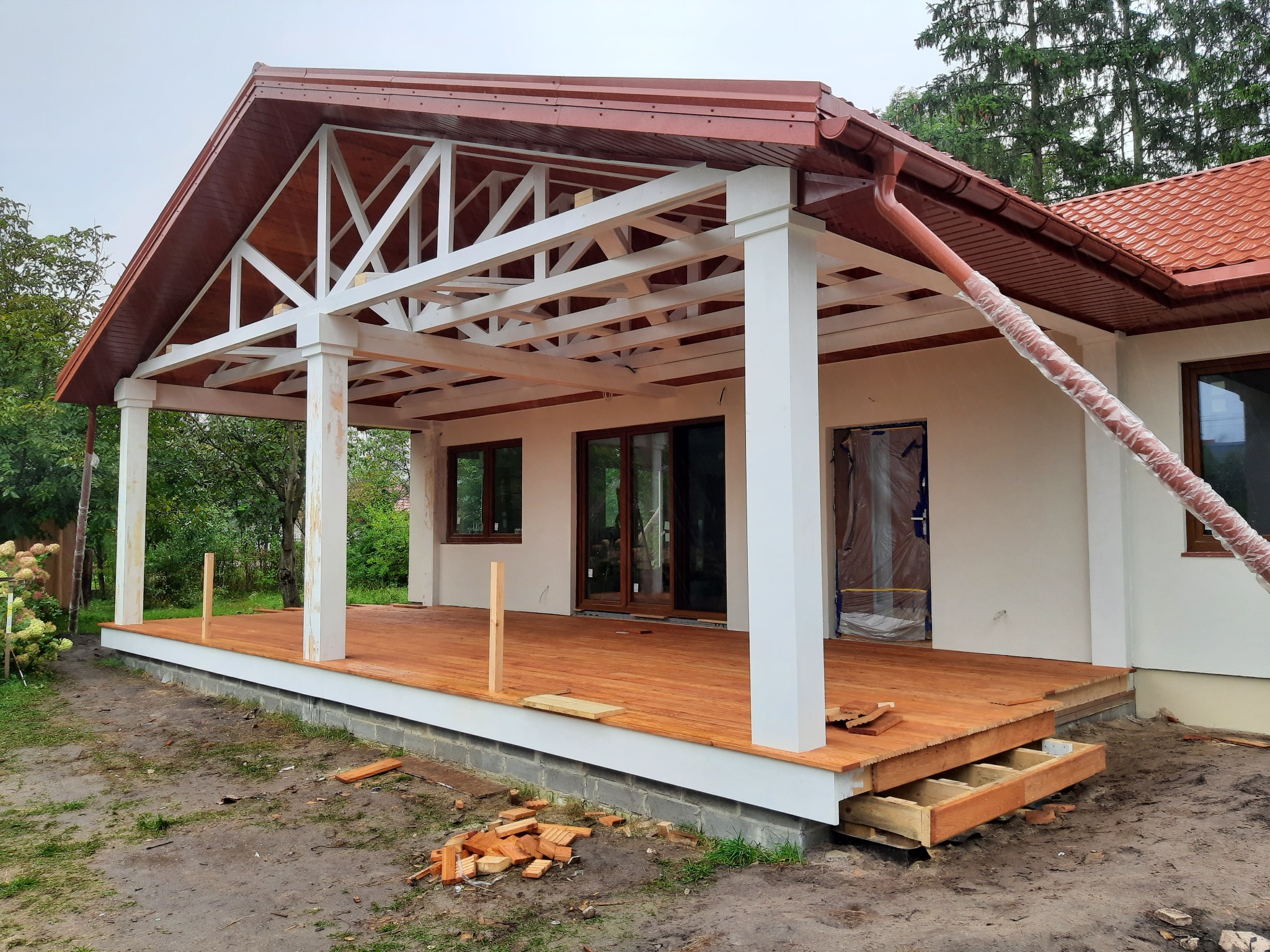 Budowa domu: widok na drewniany taras z białymi kolumnami i konstrukcją dachu, z elementami wykończeniowymi i fragmentami cegieł na ziemi.