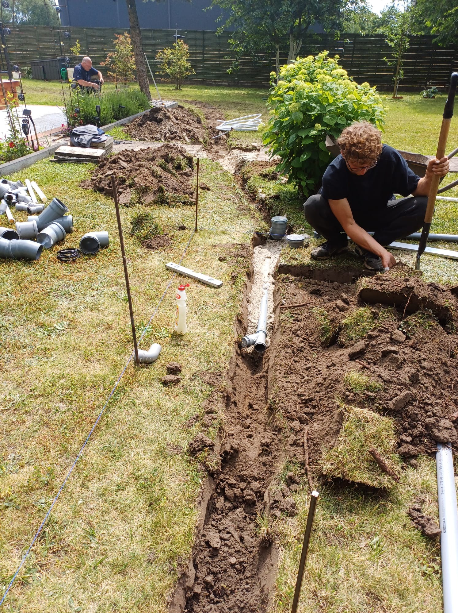 Instalacja rur kanalizacyjnych w wykopie w ogrodzie, widoczne rury, szpadel i mężczyzna w trakcie pracy, drugi mężczyzna obserwuje z tyłu.
