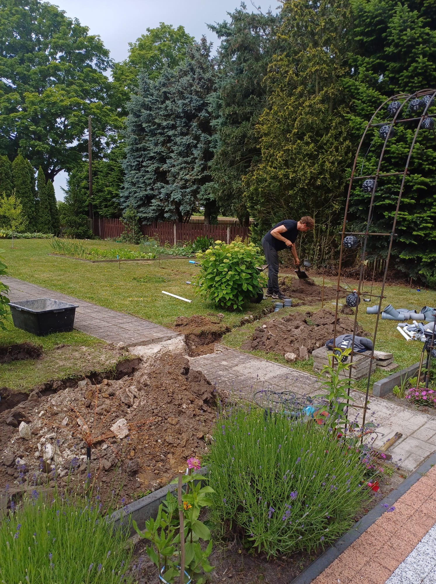 Mężczyzna kopie dół w ziemi w ogrodzie, widoczne wykopane fragmenty chodnika, rury PCV i metalowa konstrukcja pod pnącza.