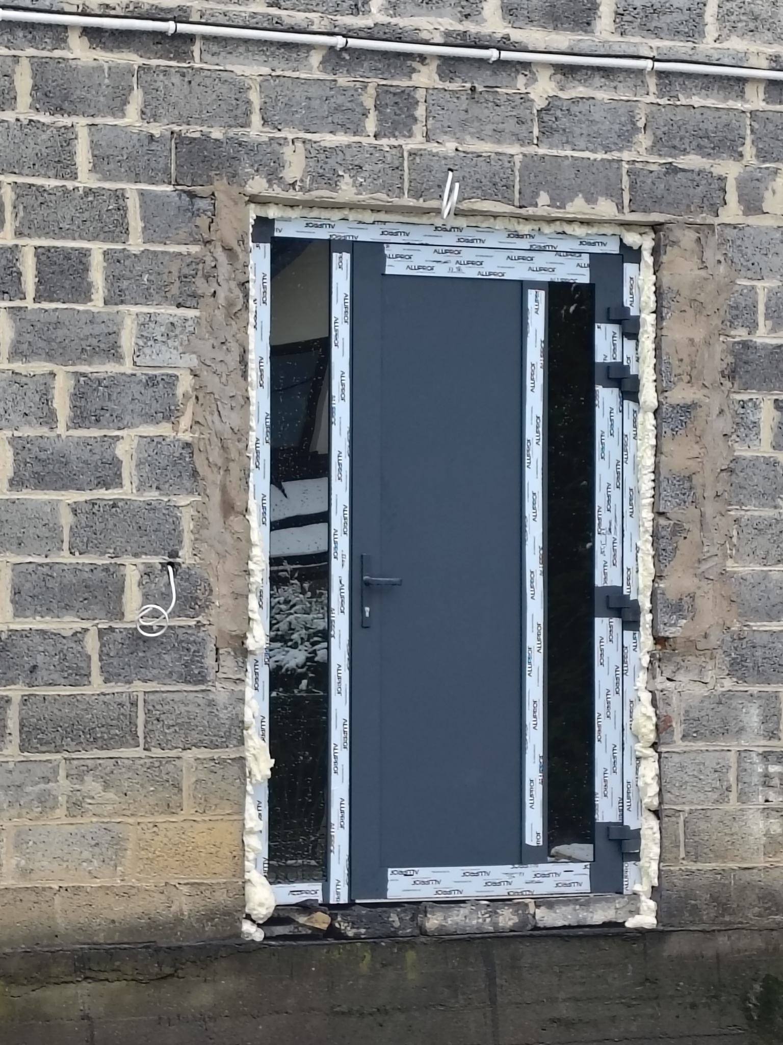 Szara metalowa ościeżnica drzwiowa z pianką montażową, osadzona w surowej ścianie z szarego bloczka betonowego. Widoczne przewody elektryczne na ścianie.