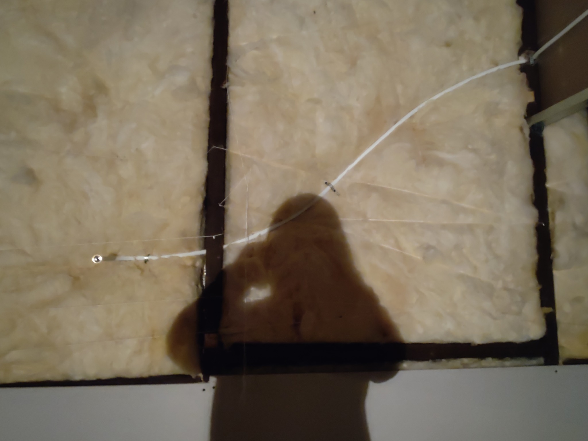 Fragment ściany z izolacją z wełny mineralnej między profilami, widoczny biały kabel mocowany do profili, na pierwszym planie cień osoby fotografującej.