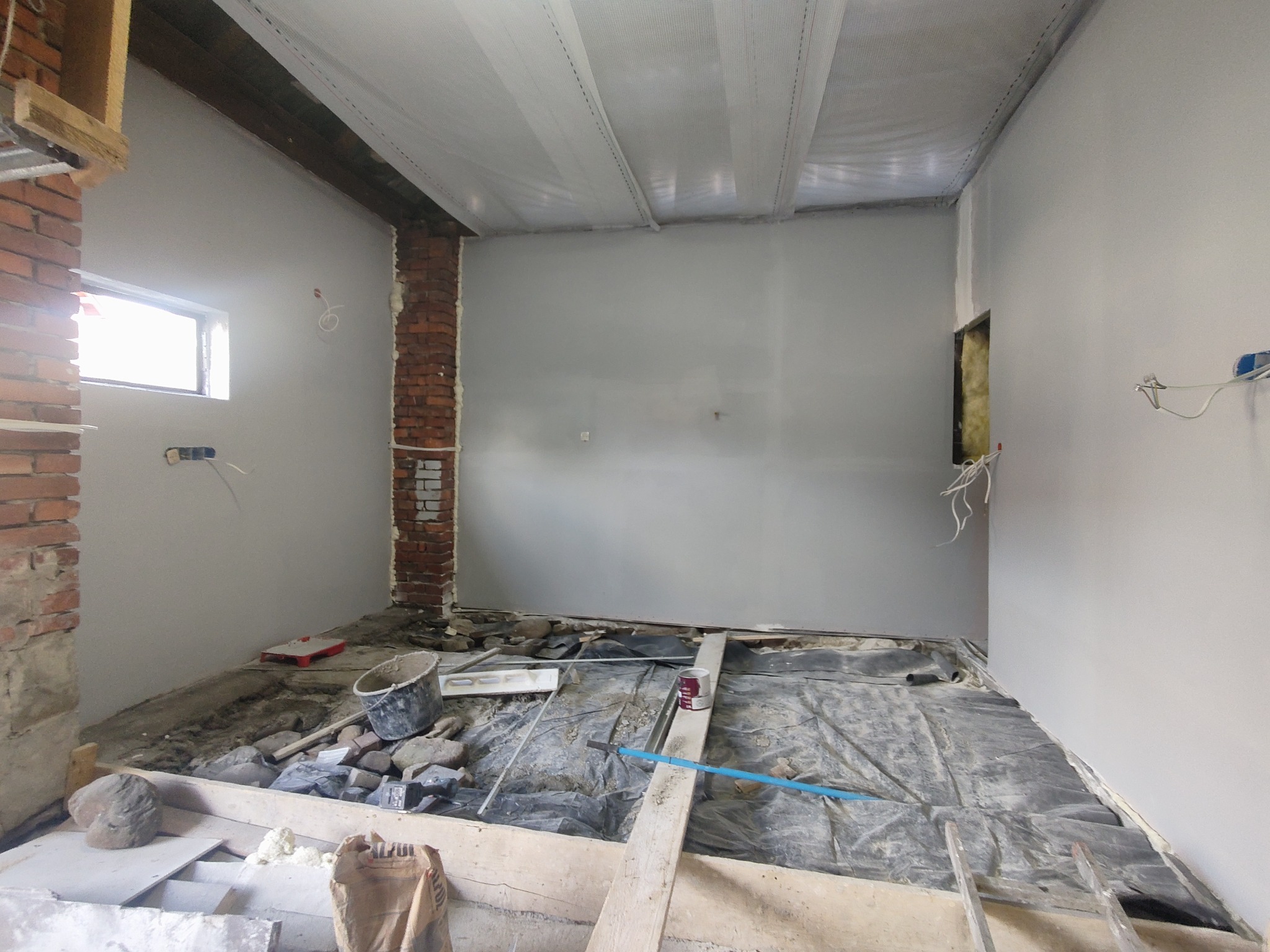 Pokój w trakcie remontu: ściany pomalowane na szaro, odsłonięta cegła, zabezpieczona folią podłoga, widoczne narzędzia i materiały budowlane, sufit zabezpieczony folią malarską.