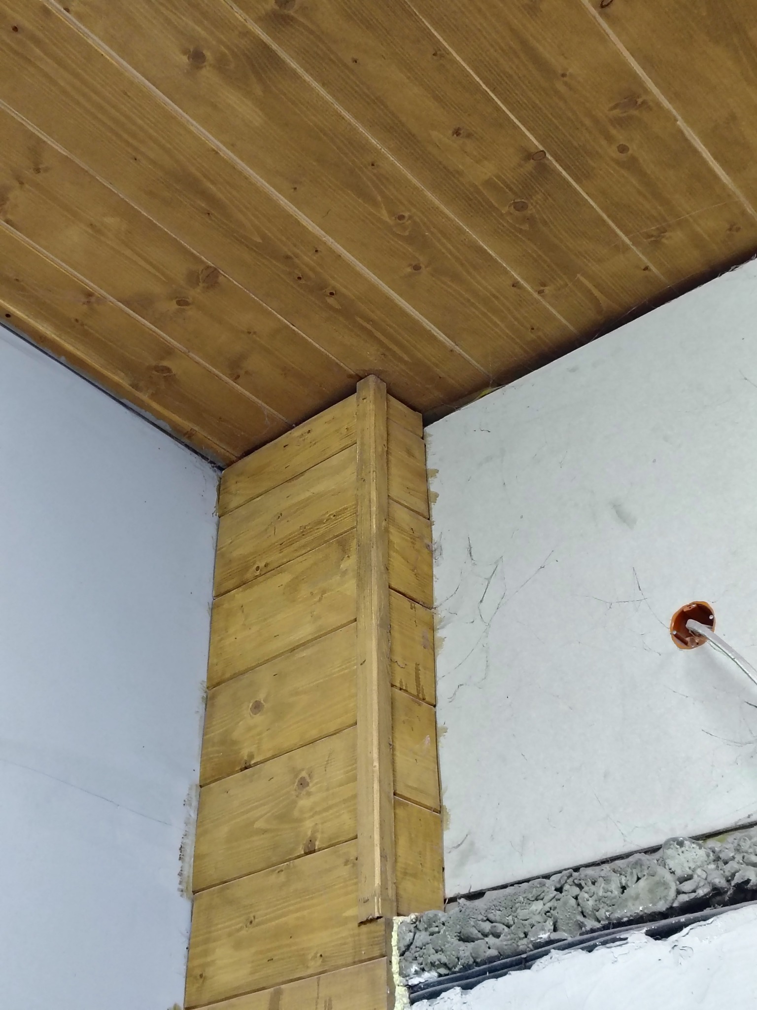 Fragment wnętrza w trakcie prac wykończeniowych: drewniana boazeria na suficie i narożniku ściany, surowa ściana z widoczną instalacją elektryczną i nierównościami w dolnej części.