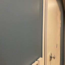 DI-NERO DMYTRO KUTSYI - Świeżo pomalowana ściana w odcieniu szaro-niebieskim, sąsiadująca z białymi drzwiami z metalową klamką i ramą. Widoczny fragment dekoracji ściennej w dolnym rogu.