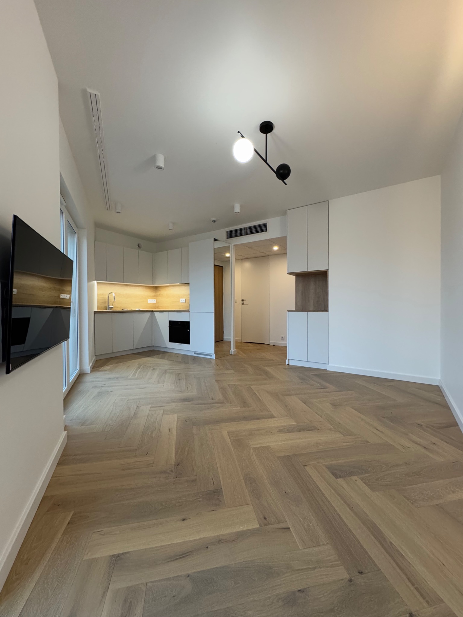 Jasne, przestronne wnętrze mieszkania z parkietem ułożonym w jodełkę, nowoczesną kuchnią z drewnianym oświetleniem i minimalistyczną lampą sufitową.