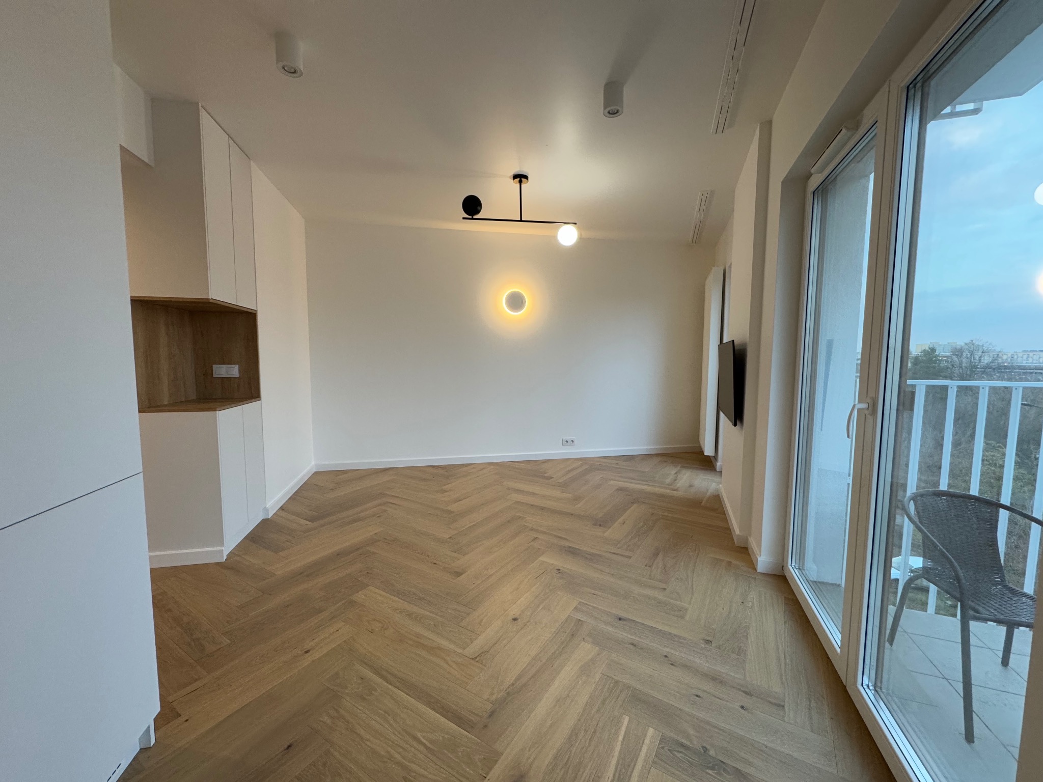 Jasne wnętrze mieszkania z parkietem ułożonym we wzór jodełki, minimalistyczną zabudową kuchenną i balkonem z widokiem na zewnątrz.