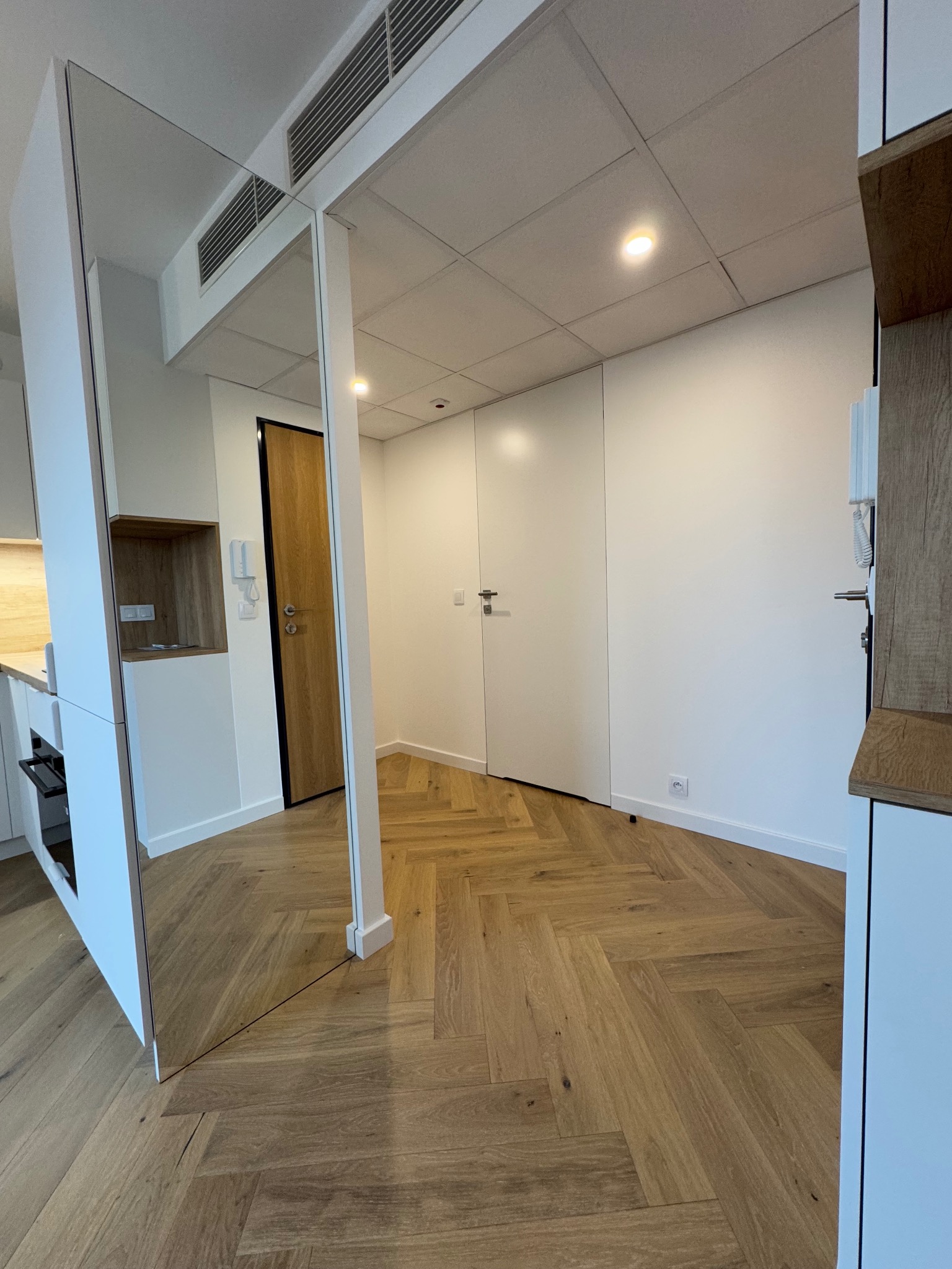 Jasne wnętrze z parkietem ułożonym w jodełkę, widok na białe ściany, drzwi i zabudowę kuchenną z elementami drewnopodobnymi.