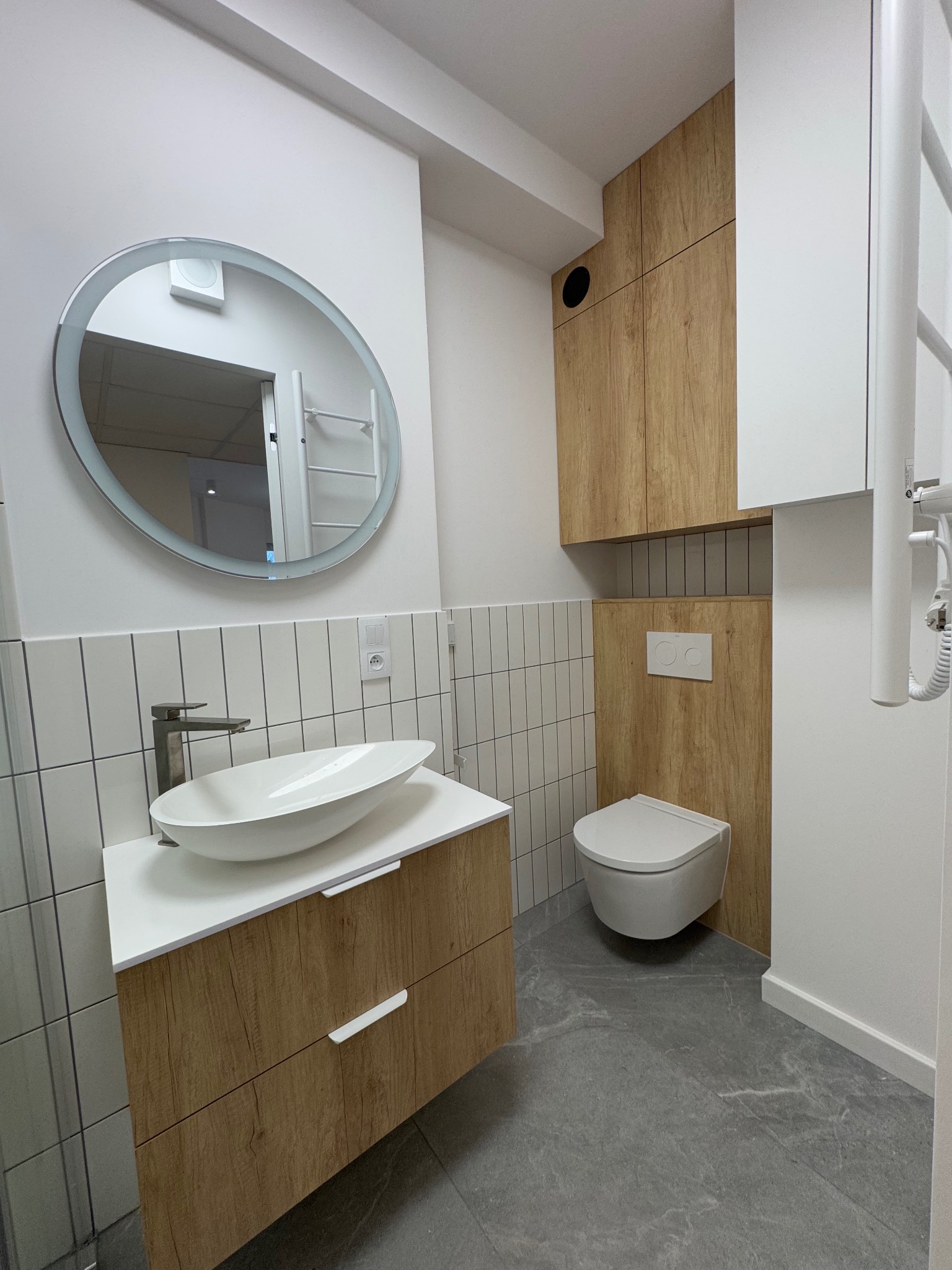 Nowoczesna łazienka z owalną umywalką nablatową, baterią w kolorze stali, okrągłym lustrem z podświetleniem LED, zabudową meblową z drewnopodobnym fornirem i szarą podłogą z płytek imitujących kamień.