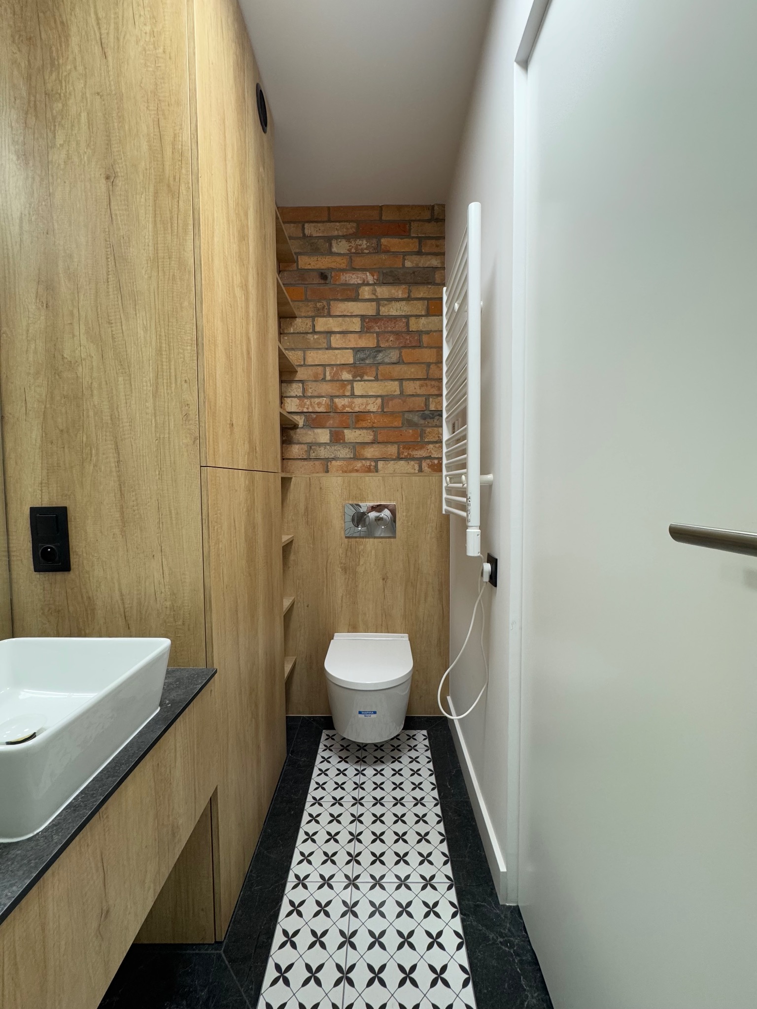 Wnętrze wykończonej łazienki z toaletą podwieszaną, umywalką nablatową, grzejnikiem drabinkowym, zabudową meblową z półkami i ścianą z cegły dekoracyjnej.