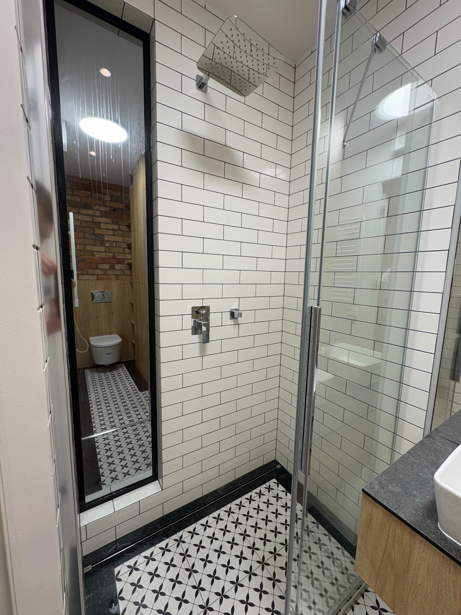 Nowoczesna łazienka z kabiną prysznicową typu walk-in, wykończona białymi, podłużnymi płytkami i czarno-białą mozaiką na podłodze, widok na toaletę w oddzielnej wnęce z ceglaną ścianą.