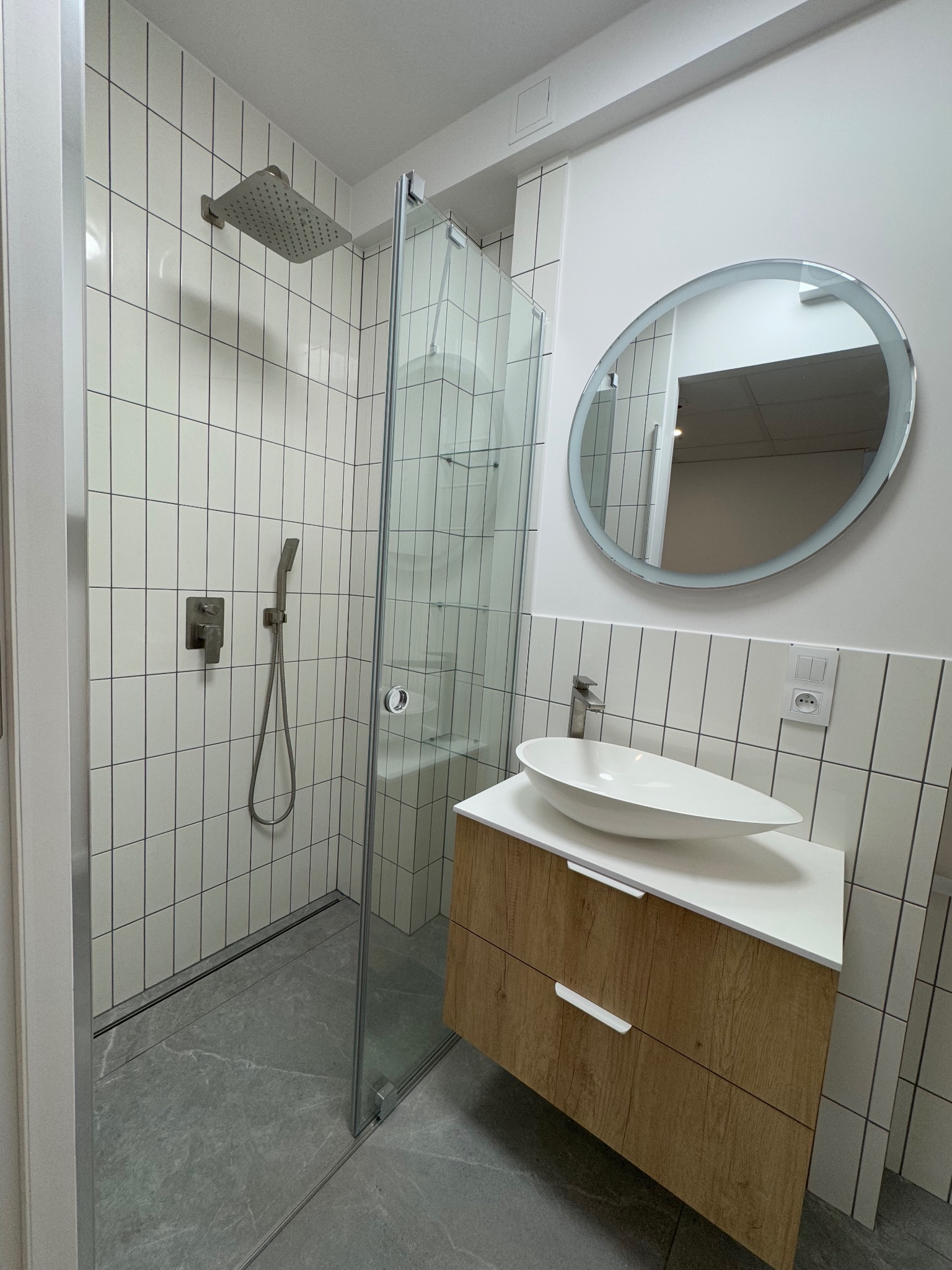 Nowoczesna łazienka z kabiną prysznicową typu walk-in, umywalką nablatową o owalnym kształcie i okrągłym lustrem z podświetleniem LED.