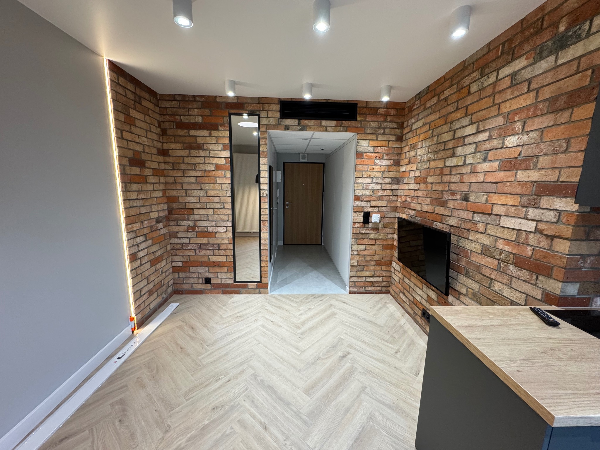 Wnętrze pokoju z ceglanymi ścianami, panelami ułożonymi w jodełkę, oświetleniem LED przy ścianie i dużym lustrem w czarnej ramie.