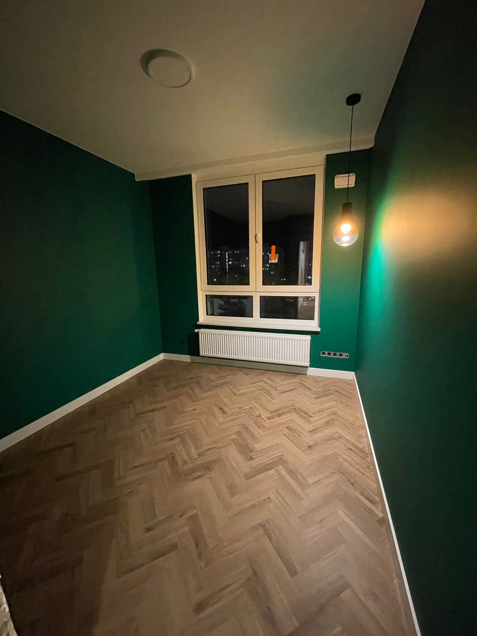 Pokój po remoncie: ciemnozielone ściany, parkiet ułożony w jodełkę, białe okno z widokiem na nocne miasto, minimalistyczna lampa wisząca z żarówką Edison.