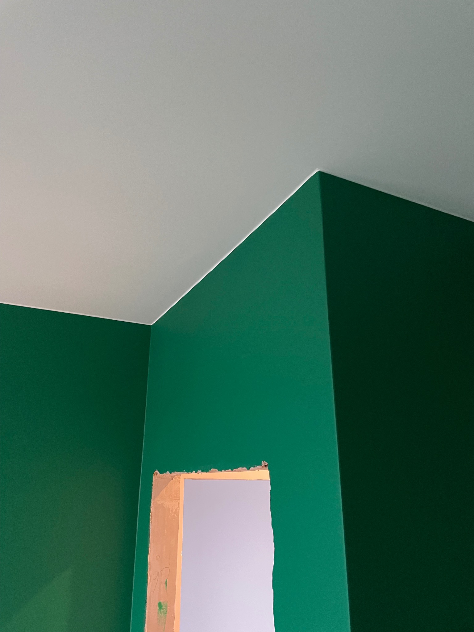 Pomieszczenie po malowaniu ścian na intensywny zielony kolor, z widocznym fragmentem surowej ościeżnicy drzwiowej i białym sufitem.