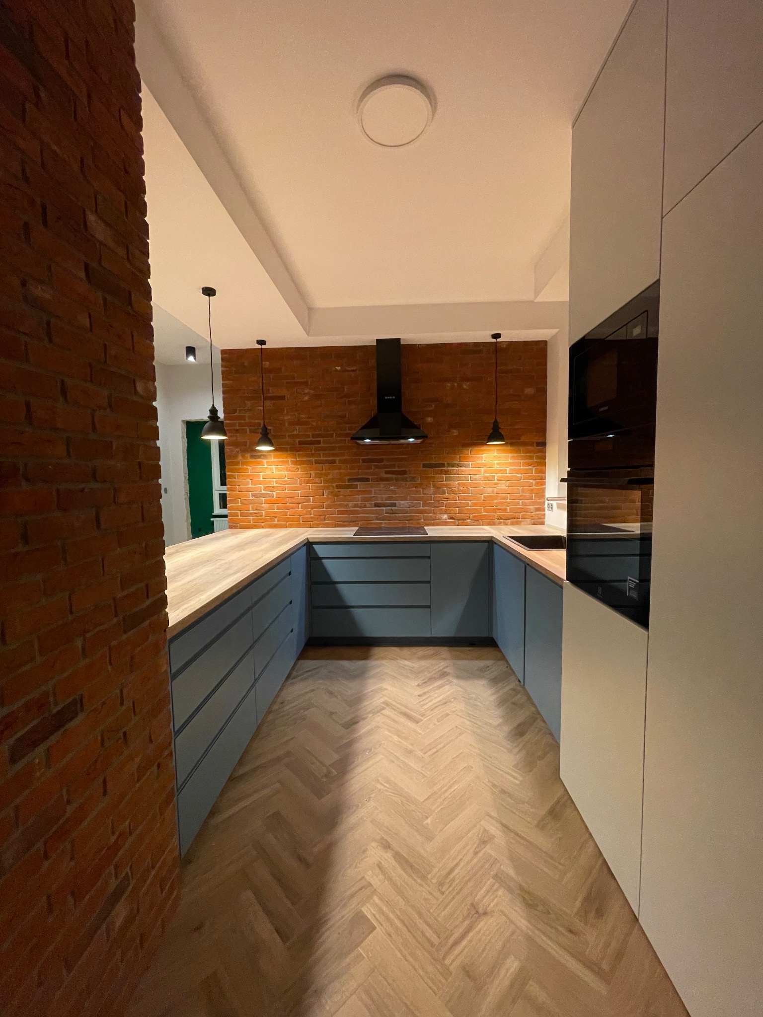 Nowoczesna kuchnia w stylu loft z ceglaną ścianą, drewnianym blatem i parkietem w jodełkę, oświetlona wiszącymi lampami.