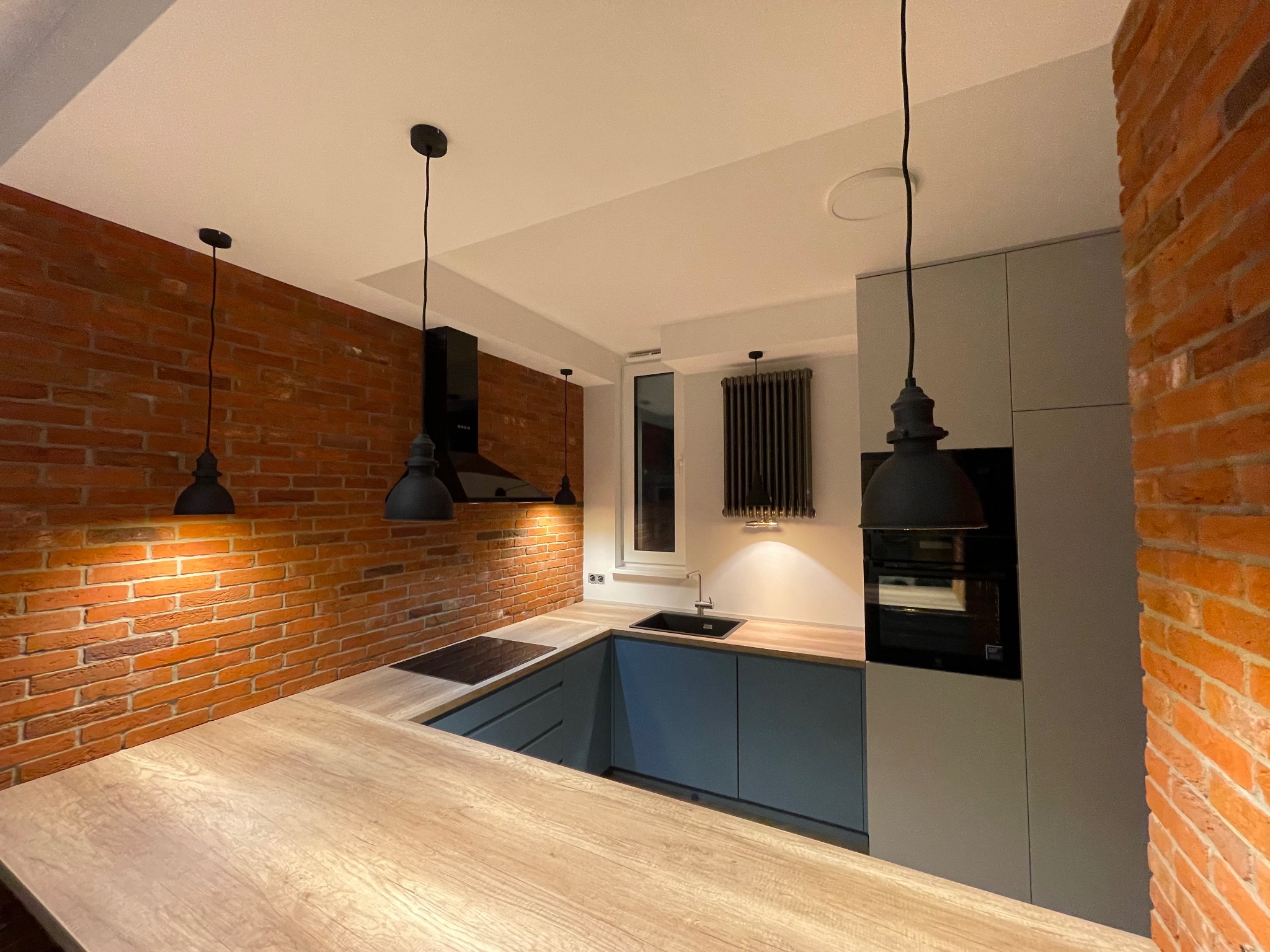 Nowoczesna kuchnia z ceglaną ścianą, drewnianym blatem, niebieskimi szafkami i czarnymi lampami wiszącymi.