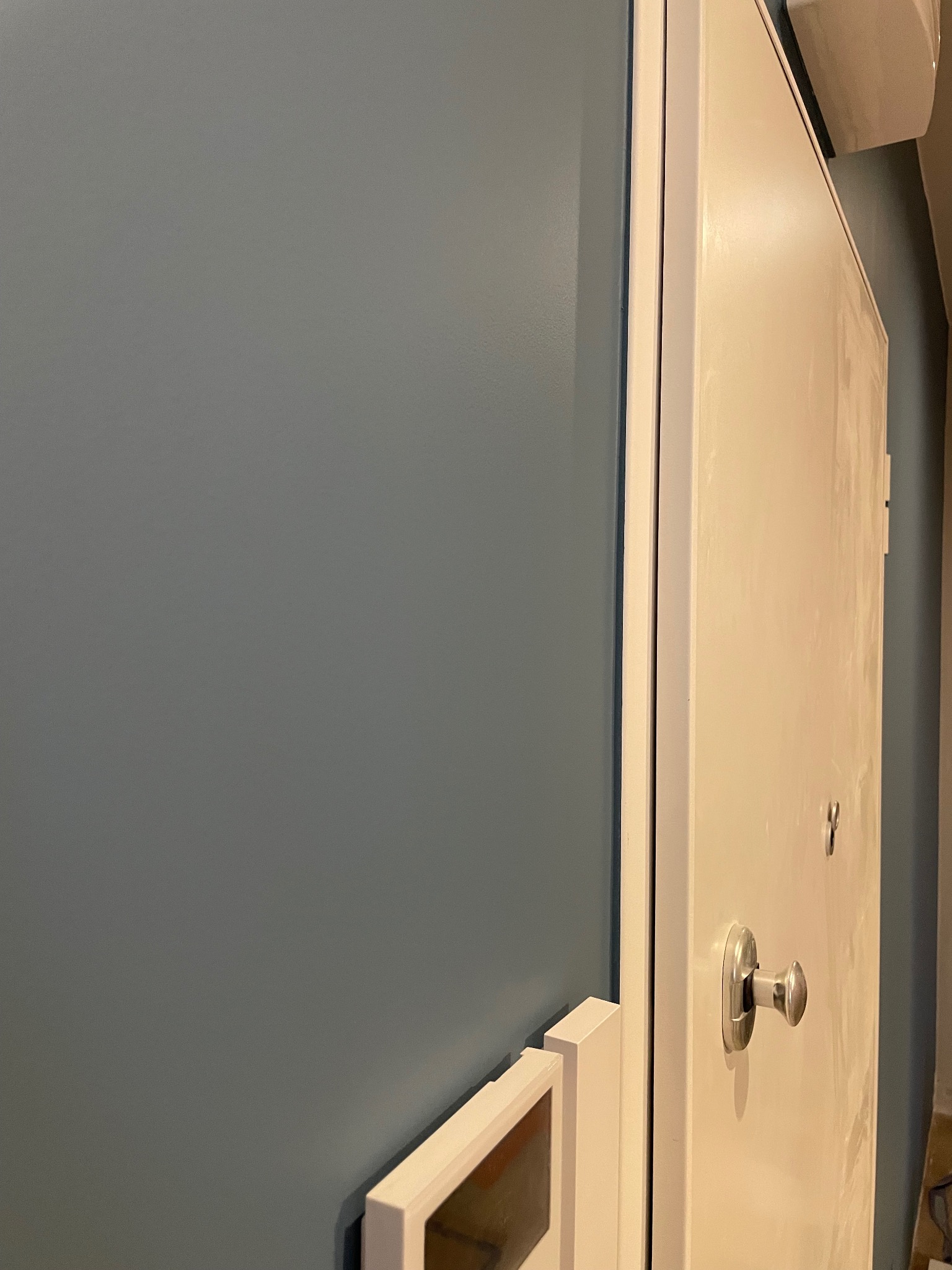 Świeżo pomalowana ściana w odcieniu szaro-niebieskim, sąsiadująca z białymi drzwiami z metalową klamką i ramą. Widoczny fragment dekoracji ściennej w dolnym rogu.