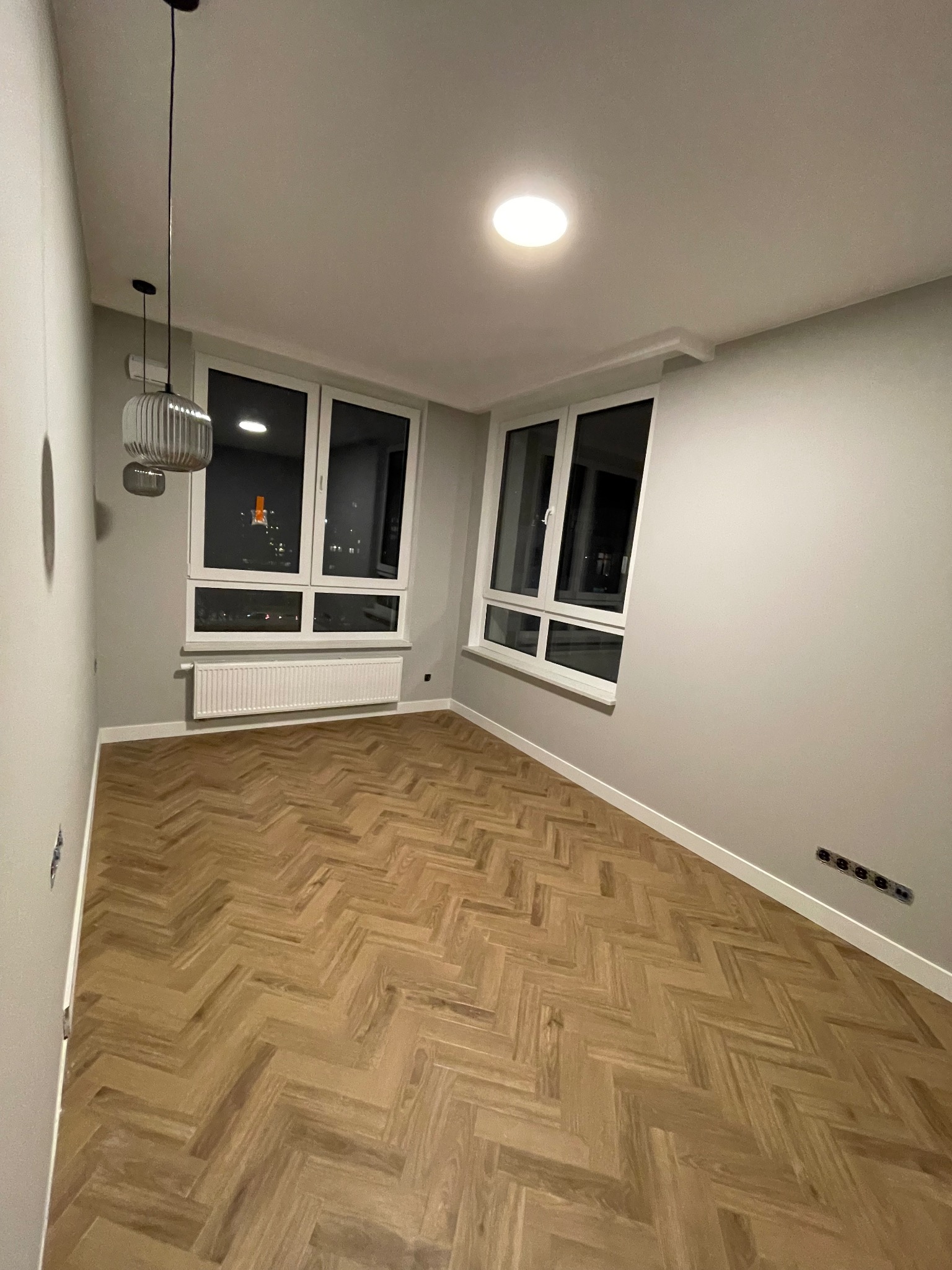 Nowoczesne wnętrze pokoju po remoncie, z parkietem ułożonym w jodełkę, świeżo pomalowanymi ścianami w odcieniach szarości, białymi listwami przypodłogowymi i grzejnikiem pod oknem.