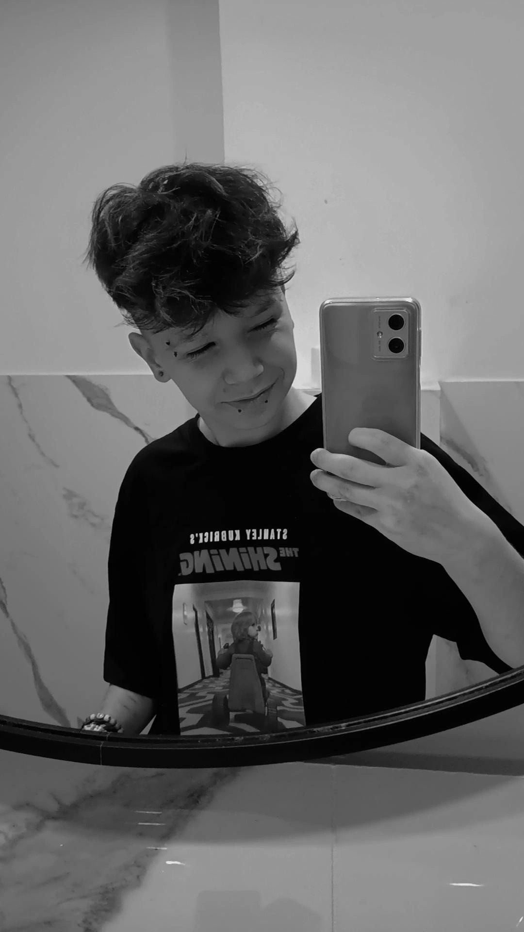 Osoba robiąca selfie w łazience, widoczna marmurowa ściana i fragment lustra, na koszulce nadruk nawiązujący do filmu 'The Shining'.