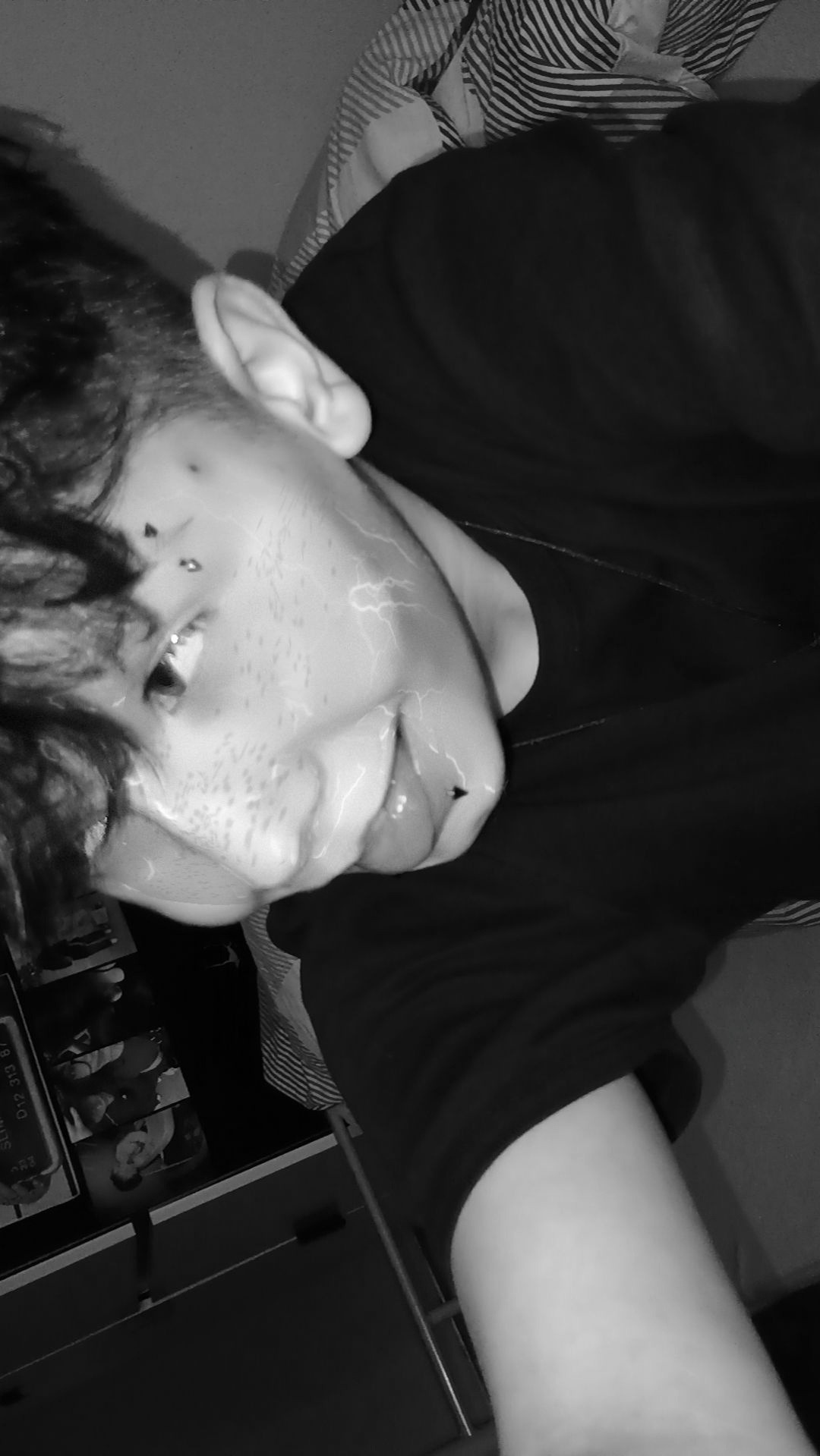 Czarno-białe zdjęcie portretowe osoby z piercingiem na twarzy, leżącej na łóżku z pościelą w paski, z widocznymi zdjęciami na szafce obok.